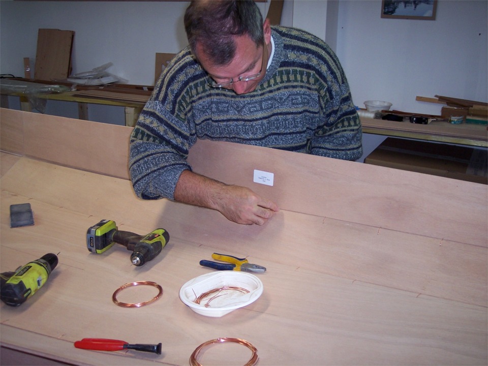 Gilles enchaîne avec le bordé 2 tribord. Nous avons préalablement percé les trous le long du bord haut du bordé 1, il s'agit de percer le bordé 2 en regard de chaque trou de suture dans le bordé 1, puis de passer une suture en fil de cuivre dans chaque trou, afin de lier les bordés entre eux, puis de les coller.