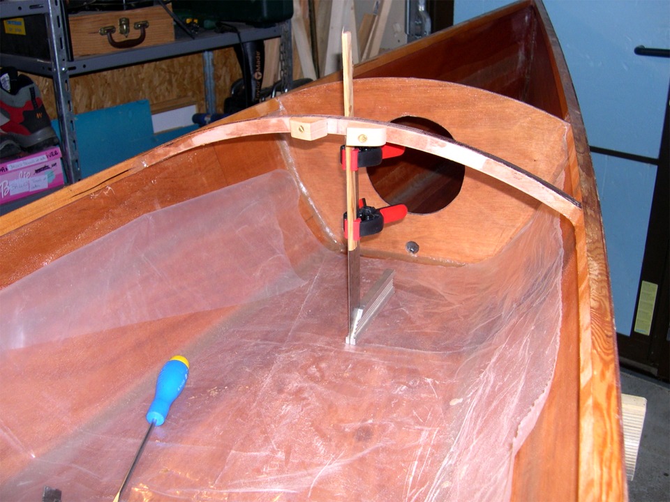 Tous les éléments précités ont été collés et Olivier est en train de positionner le barrot de l'avant du cockpit qui recevra l'avant des élongis entre les deux pièces triangulaires. La courbe du barrot montre que le pont avant a un bouge assez prononcé afin d'augmenter le volume de la partie avant de la coque : donc plus de réserve de flottabilité et plus de place pour les jambes du kayakiste avant, ainsi que pour les bagages. 