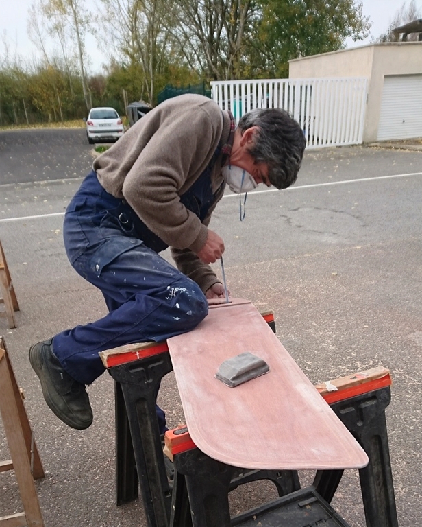 Ce matin, le ponçage rituel est assez technique, comme le montre la position de Pierre, qui cale la dérive avec son genou tandis qu'il ponce l'intérieur de la poignée à l'aide d'un lacet découpé dans une garniture de ponceuse à bande au rebut.