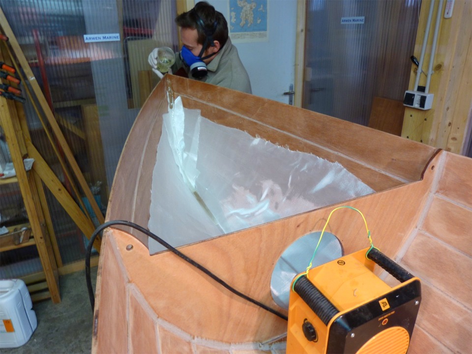 Arnaud est revenu pour faire la stratification sur l'intérieur de la Yole de Chester, un peu négligée ces temps-ci. Nous commençons par le compartiment étanche avant, qui est la partie la plus compliquée, du fait de la verticalité des parois et du manque d'accessibilité dans l'étrave, où il y a justement trois couches de tissu de verre à saturer. Arnaud verse l'époxy dans l'étrave : elle va s'accumuler dans le brion, puis se répandre le long de la ligne de quille, en ayant mouillé au passage le tissu au creux de l'étrave, ce qui facilitera un peu sa saturation à la squeegee (et au pinceau pour les zones trop inaccessibles). Le radiateur soufflant aide à préchauffer le compartiment central (il faisait -11°C ce matin à Clairoix). 