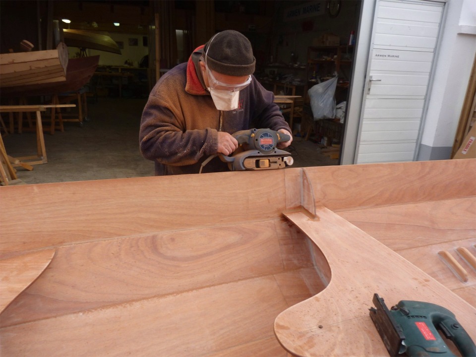 Derni&egrave;re op&eacute;ration de cette construction accompagn&eacute;e : le fa&ccedil;onnage des listons. Nous avons commenc&eacute; par passer un coup de "tank" (ponceuse &agrave; bande) sur la face inf&eacute;rieure des listons afin d'&eacute;liminer les exc&eacute;dents d'&eacute;poxy durcie et mettre &agrave; niveau les deux lattes, puis nous passons le "tank" sur la face sup&eacute;rieure (ce que fait Herv&eacute; sur cette image), et nous finissons par d&eacute;couper les extr&eacute;mit&eacute;s en arrondi, puis par passer un coup de d&eacute;fonceuse afin d'arrondir leur ar&ecirc;te sup&eacute;rieure ext&eacute;rieure. Herv&eacute; devra encore coller le skeg et passer une couche d'&eacute;poxy sur toute la surface int&eacute;rieure et ext&eacute;rieure. 