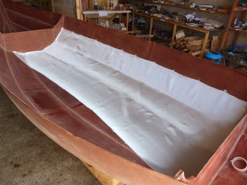 J'ai encore poncé tout l'intérieur de la coque, et je l'ai dépoussièré afin d'éviter qu'il reste des poussières sous le tissu de verre (qui seraient cause de bulles). Je viens de draper la première des trois pièces de tissu qui vont se chevaucher pour couvrir la sole et les bordés 1 et 2. Seuls les bordés 3 ne seront pas stratifiés sur l'intérieur. Le tissu couvre leur joint avec les bordés 2 pour renforcer celui-ci, mais il y aurait peu d'intérêt structurel à couvrir aussi l'intérieur des carreaux, et cela ajouterait encore près d'un kilo de poids, localisé en haut de la coque qui plus est. 