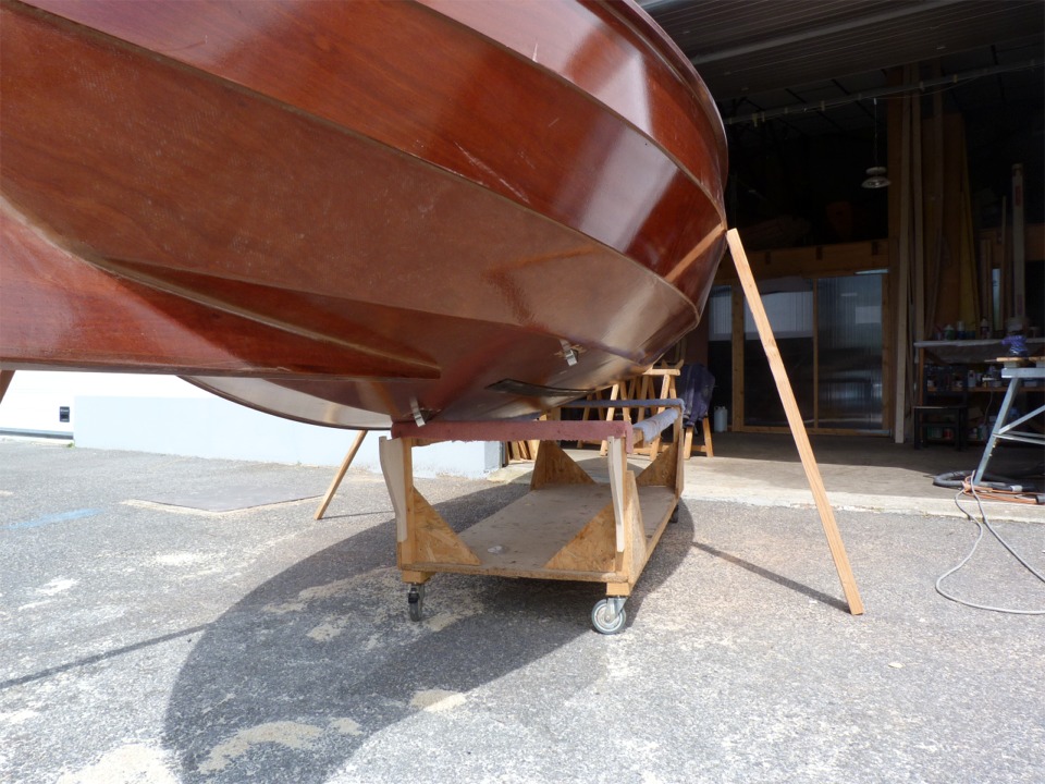 J'utilise des béquilles sommaires afin de pencher la coque pour monter les vide-vite, que l'on aperçoit, en place et ouverts tous deux. Leur face arrière verticale génère une aspiration du fait du déplacement de la carène dans l'eau. Un clapet est alors ouvert par l'effet de succion (effet venturi) et l'eau des fonds est aspirée à l'extérieur, ce qui évite l'écopage obligatoire par bonne brise sur un bateau ouvert. Malheureusement, les essais au lac d'Orient ont montré que l'effet venturi n'est pas au rendez-vous car l'emplacement des vide-vite n'est pas bon : sans doute trop proche de l'angle extérieur de la sole. Je vais donc les démonter, boucher les trous et en refaire d'autres à un endroit plus proche de l'axe de la coque. 
