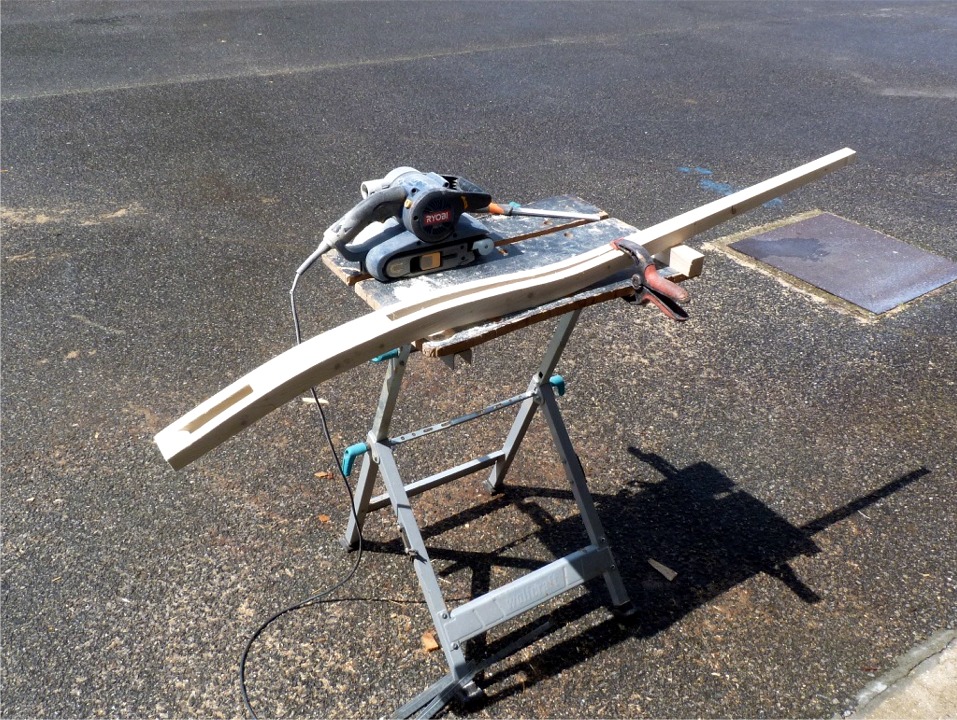 Je finis d'ajuster la découpe de la barre de mon Grand Skerry "Gandalf" à la ponceuse à bande. J'ai découpé deux demi-barres en forme de S très aplati dans une planche de pin de 20 mm, les ai collées autour d'une "âme" du même métal en les affinant coté poignée pour créer un long biseau de collage. Deux tiges filetées de laiton avec écrous noyés renforcent le collage de part et d'autre de la mortaise qui chevauchera le tête du gouvernail. 