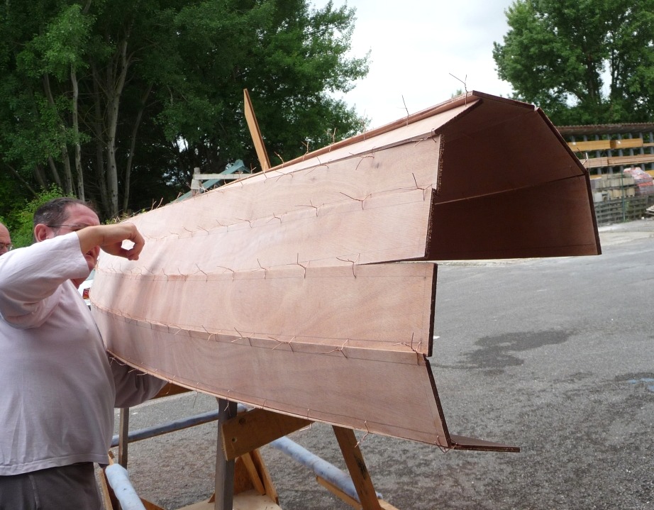 L'averse est pass&eacute;e et nous sommes de nouveau dehors. Emmanuel passe le bras sous la partie tribord de la coque afin de la relever et d'y accrocher une "b&eacute;quille" afin de la soutenir. Le bord&eacute; 5 est en place du cot&eacute; b&acirc;bord, alors qu'il n'y en a encore que 4 sur tribord. 