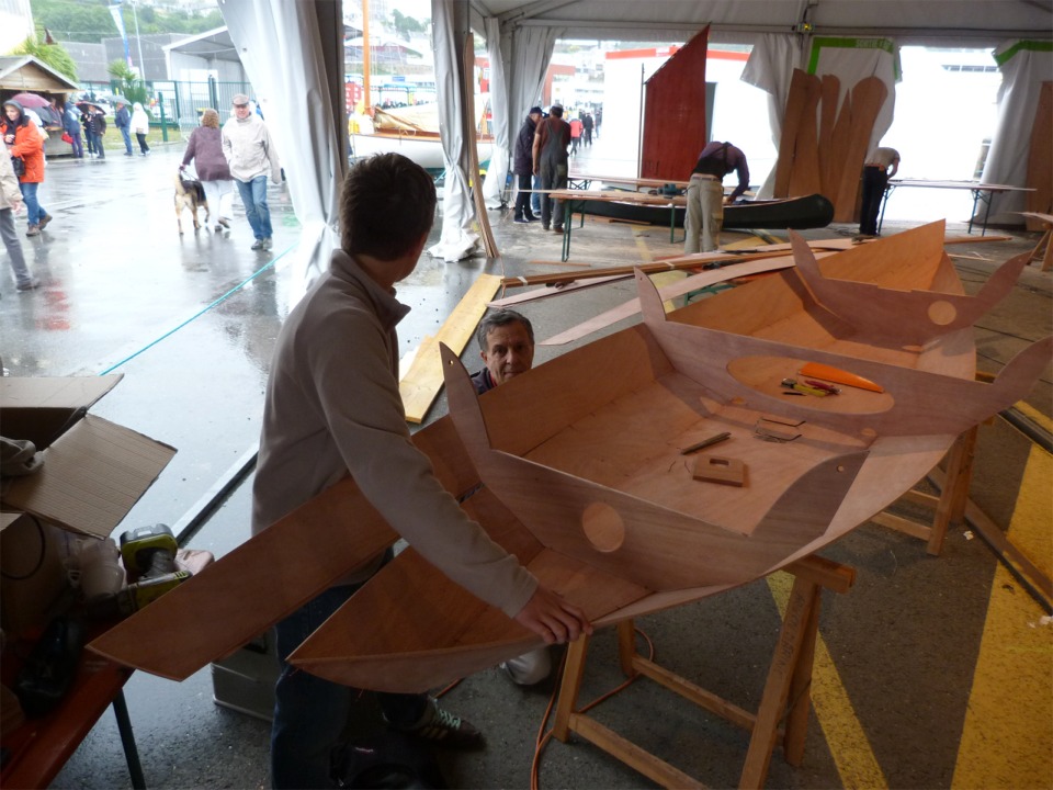 Michel et François ont en mis place les trois membrures et procèdent au montage du bordé 2 bâbord. On aperçoit à l'arrière-plan l'équipe de l'Atelier Bois Courbe et le Caïman "modèle" avec sa petite voile aurique. 