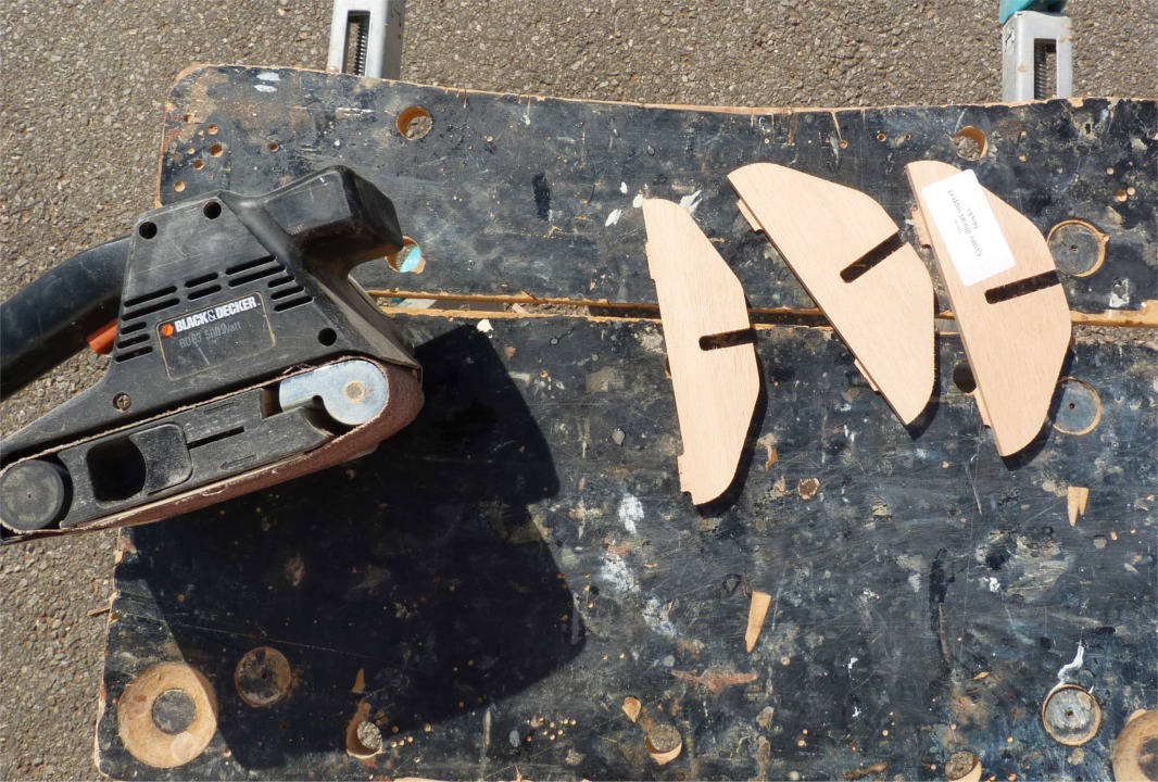 Voici les trois petits supports du banc central, qui s'encastrent dans les trois fentes du haut de la membrure centrale. Au pr&eacute;alable, je vais supprimer &agrave; la ponceuse &agrave; bande les ergots laiss&eacute;s par la machine de d&eacute;coupe num&eacute;rique. Ces ergots servent &agrave; conserver une coh&eacute;sion minimale des pi&egrave;ces dans la feuille de contreplaqu&eacute;, afin de faciliter l'extraction des pi&egrave;ces apr&egrave;s la d&eacute;coupe. 
