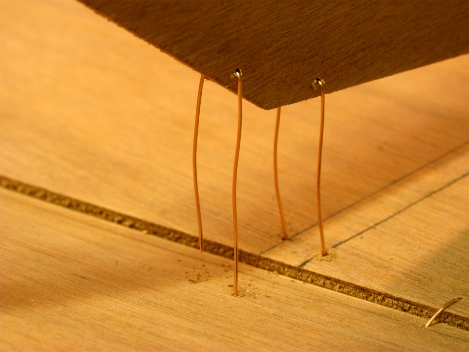 Gros plan sur les deux sutures de l'angle inf&eacute;rieur tribord de la cloison avant. J'ai ins&eacute;r&eacute; d'abord les sutures dans les trous perc&eacute;s dans la cloison, je les ai pli&eacute;es en "U", et j'ai ins&eacute;r&eacute; chacune des pointes des "U" dans les deux trous perc&eacute;s &agrave; travers la sole. On voit d'ailleurs que l'extr&eacute;mit&eacute; la plus &agrave; gauche est plus courte n'est pas encore entr&eacute;e dans son trou. Notez que l'on voit la tranche de la sole en surplomb par rapport au bord&eacute;. En effet, la sole est r&eacute;alis&eacute;e en contreplaqu&eacute; de 9 mm d'&eacute;paisseur, tout comme les cloisons, alors que les bord&eacute;s sont en 6 mm. On aper&ccedil;oit une suture joignant le bord&eacute; &agrave; la sole au premier plan &agrave; droite.