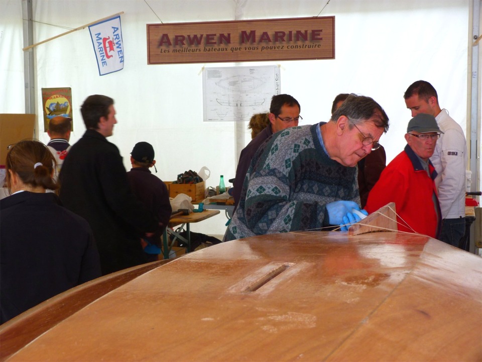 Nous enchainons avec le collage de l'aileron : nous commençons par encoller son chant, puis nous le positionnons sur le tracé que j'ai fait lors de la préparation. Nous vérifions qu'il est bien orthogonal par rapport à la sole, puis nous le fixons avec du scotch (enfin pas exactement, c'est de la bande à masquer qui le maintient en place en attendant que l'époxy durcisse). 