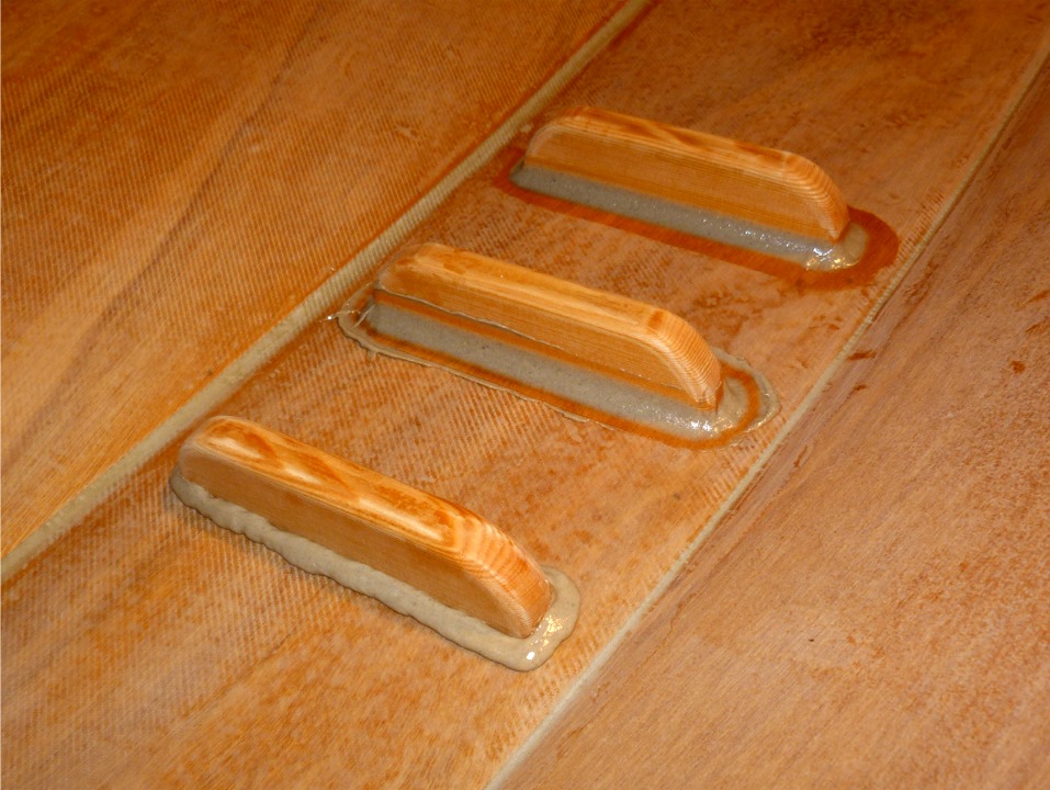 Apr&egrave;s avoir fix&eacute; les cale-pieds, je fais de petits joints-cong&eacute;s autour de leur base afin d'am&eacute;liorer leur tenue en vue des efforts qu'ils seront appel&eacute;s &agrave; subir. Ce gros plan montre les trois &eacute;tapes de la confection de ces joints-cong&eacute;s. On commence (&agrave; gauche) par d&eacute;poser &agrave; la poche &agrave; douille ou &agrave; la seringue un cordon d'&eacute;poxy charg&eacute;e de consistance "cr&egrave;me de marrons" et de diam&egrave;tre ad&eacute;quat, puis (milieu) on forme le joint avec le dos d'une cuiller, le coin arrondi d'une chute de contreplaqu&eacute; ou tout autre outil ad hoc, et on finit en ramassant l'exc&eacute;dent d'&eacute;poxy sur les bords du joint-cong&eacute; &agrave; l'aide d'un couteau &agrave; mastic. 