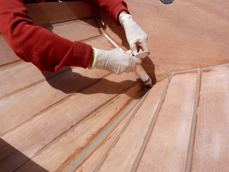 Annick utilise une seringue pour déposer un filet d'époxy chargée sur le joint entre les bordés 1 et 2 au pied du tableau. Notez l'importance du joint sur la ligne de quille à l'intérieur du caisson arrière. 