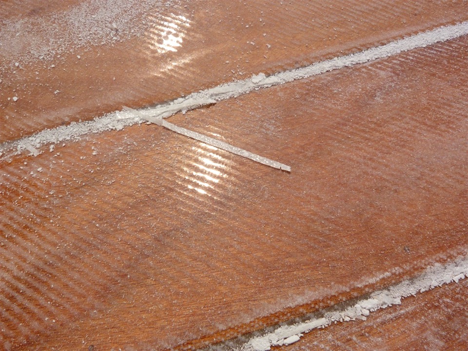 Gros plan sur la surface stratifi&eacute;e apr&egrave;s pon&ccedil;age : la poudre blanche est surtout constitu&eacute;e d'&eacute;poxy, mais il y a aussi un peu de fibre de verre, parfois en morceaux assez gros, comme la baguette que l'on voit au centre le l'image, qui fait 7 cm de long. Ce morceau provient de la lisi&egrave;re de la stratification, sur le joint entre les virures 2 et 3. Par endroits, le bord du tissu s'est l&eacute;g&egrave;rement relev&eacute; suite &agrave; la coupe, et j'&eacute;limine toute la fibre de verre qui n'est pas en contact avec le contreplaqu&eacute;, car le composite est incomplet. 