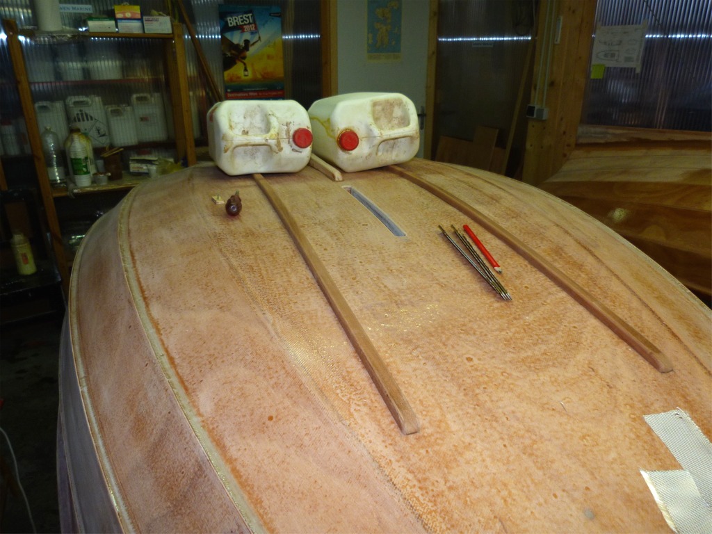 Je passe &agrave; la pr&eacute;paration du collage des fausses-quilles lat&eacute;rales de la prame. Les deux lattes de sapelli constituant ces fausses quilles sont tenues par une vis temporaire &agrave; chacune de leurs extr&eacute;mit&eacute;s pendant le durcissement de l'&eacute;poxy de leur collage. Je viens de visser la vis de l'avant, apr&egrave;s avoir fait un trou &agrave; travers la sole et un avant-trou dans le latte de sapelli, puis je presse (&agrave; l'aide des deux bidons de 10 litres) l'extr&eacute;mit&eacute; arri&egrave;re des lattes align&eacute;es &agrave; l'arri&egrave;re afin de percer l'avant-trou arri&egrave;re, puis visser la vis arri&egrave;re.