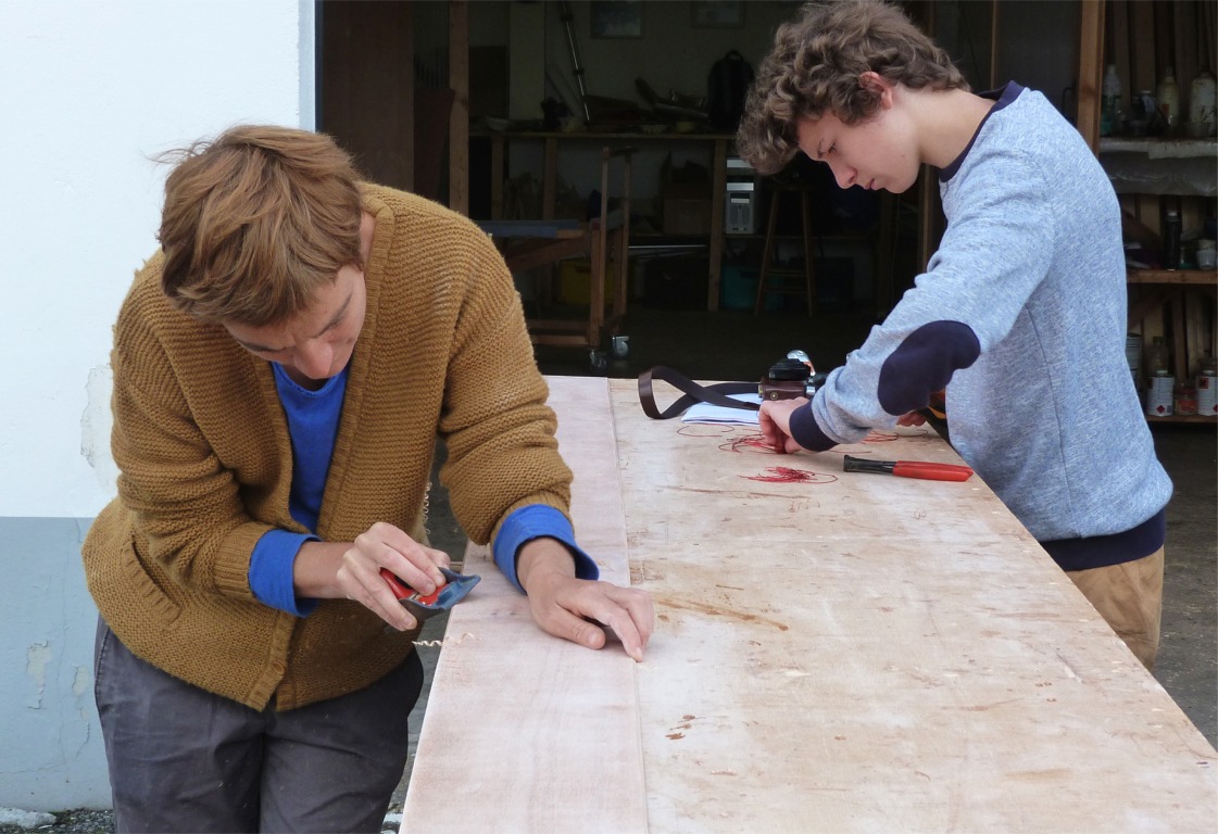 Maud et Gaël ont fait tout le chemin depuis l'Ariège pour venir construire à l'atelier un Wood Duck 12. J'ai préparé le kit en collant les joints-puzzle des demi-panneaux et en passant une couche d'imprégnation époxy sur toutes les pièces, que j'ai ensuite poncées à plat. Nous finissons ensemble la préparation : Maud fait un biseau au rabot sur les bords des panneaux afin de réduire l'ouverture des joints que nous allons faire entre les panneaux. Gaël coupe les rouleaux de fil de cuivre en segments de 8 à 10 centimètres que nous allons utiliser pour coudre les panneaux entre eux. 