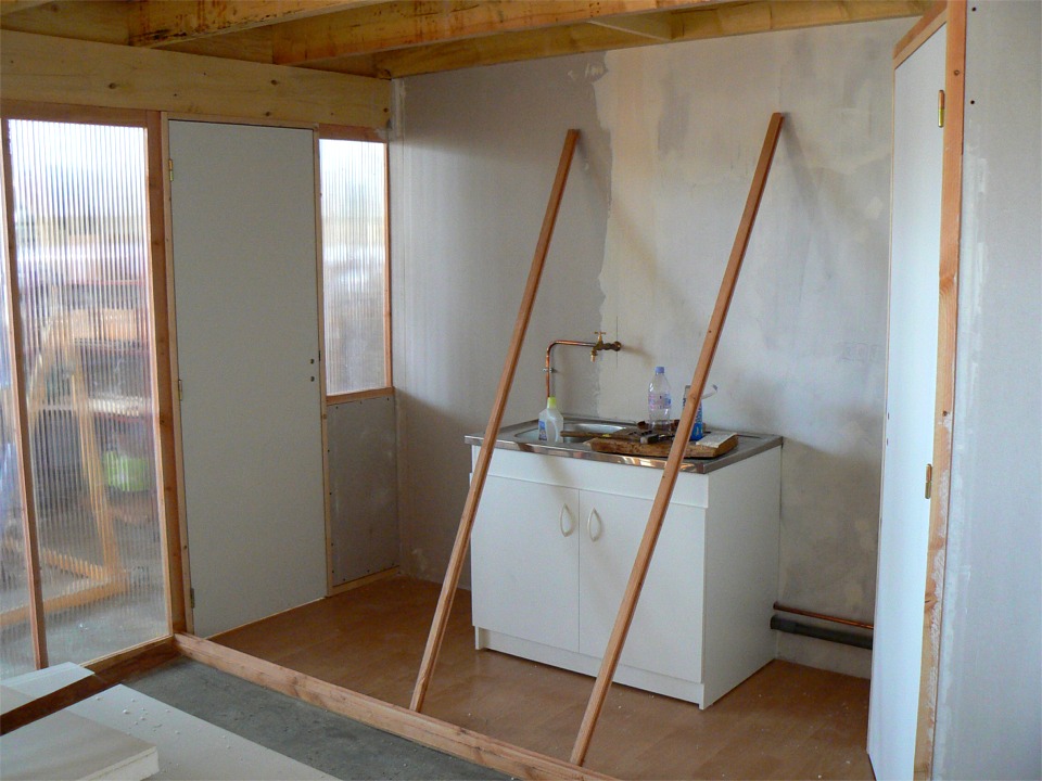 Un peu plus tard, les toilettes sont ferm&eacute;es et la cloison de fa&ccedil;ade ext&eacute;rieure de la "cuisine" est en place. Les deux tasseaux appuy&eacute;s sur l'&eacute;vier ont &eacute;t&eacute; rainur&eacute;s &agrave; la d&eacute;fonceuse pour tenir la cloison entre la cuisine et "l'atelier couvert" sous la mezzanine. Cette cloison sera faite de plaques de polycarbonate cloisonn&eacute; en trois &eacute;paisseurs : translucide (oui je sais, extralucide, c'est autre chose) et isolant. Le pouvoir isolant de cette cloison sera suffisant car le diff&eacute;rentiel sera rarement au-dessus de 15&deg;C, et elle ne sera pas directement expos&eacute;e aux intemp&eacute;ries. Notez le luxueux parquet de r&eacute;cup', cadeau de Dominique. 