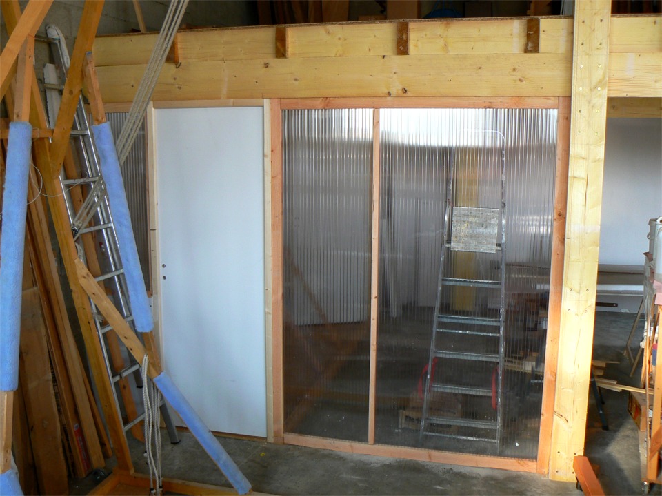 La m&ecirc;me zone est ici vue de "l'ext&eacute;rieur" : l'acc&egrave;s de la cuisine et des toilettes (par la porte &agrave; gauche) est s&eacute;par&eacute; de la partie "atelier couvert" afin que le passage ne contamine pas un vernis en cours par exemple, ou encore pour que le caf&eacute; n'ait pas un petit arri&egrave;re-go&ucirc;t de solvant... Le bout de cloison en polycarbonate &agrave; droite de la porte ferme le reste de la port&eacute;e gauche (le madrier de fa&ccedil;ade de la mezzanine est en trois port&eacute;es). 