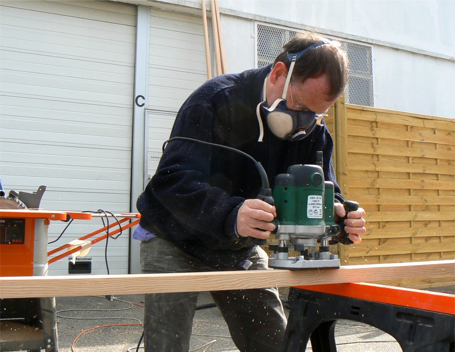 Les voilà en effet de retour à l'atelier, et nous voyons ici Olivier à la défonceuse, en train d'arrondir les angles de sa livarde. Nous avons décidé de laisser tous les espars de section essentiellement carrée, et de nous limiter à arrondir les angles. Par contre, nous avons affiné les extrémités du mât et de la livarde sur la scie que l'on aperçoit derrière Olivier. 