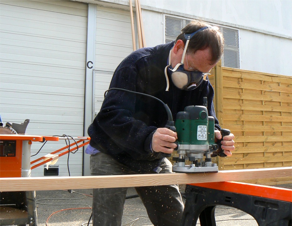 Les voilà en effet de retour à l'atelier, et nous voyons ici Olivier à la défonceuse, en train d'arrondir les angles de sa livarde. Nous avons décidé de laisser tous les espars de section essentiellement carrée, et de nous limiter à arrondir les angles. Par contre, nous avons affiné les extrémités du mât et de la livarde sur la scie que l'on aperçoit derrière Olivier. 