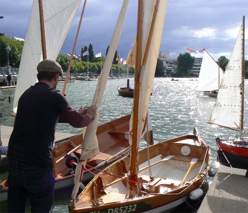 Ludo est occupé à ferler le foc de son "Anouket" pendant qu'Olivier débarque de "Malo" par l'avant, ce qui explique sa position, avec le safran totalement à l'air. La manoeuvre d'Olivier est un peu difficile à effectuer sur un bateau plus léger que lui... A l'arrière-plan, Jean-Jacques passe avec "Keltia" et Gérard affale sa voile sur "La Marie Pupuce" pour accoster à l'aviron. A droite, on aperçoit l'artimon de l'étonnant modèle réduit de "Joshua", le bateau conçu par Moitessier et avec lequel il réalisera ses plus fameuses traversées.