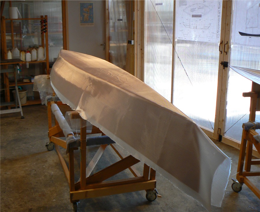 Après avoir fait quelques retouches sur les joints extérieurs et encore poncé, puis fastidueusement dépoussiéré, j'en viens enfin à la stratification de la coque : j'ai drapé le tissu de verre sur la coque, j'ai coupé le plus gros de l'excédent au livet et je suis prêt à mixer l'époxy. Je couperai l'excédent de tissu sur l'étrave, au premier plan, après qu'il soit saturé et j'ajouterai une bande de tissu à cheval sur la ligne d'étrave pour la renforcer. 