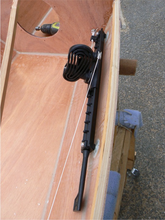 Vue de dessus : le câble est fixé à l'avant du rail, s'enroule autour du bas de la pédale, puis autour de la partie métallique en haut, solidaire de la pédale. C'est le changement d'incidence de la pédale qui tend ou détend le câble. (Hein? Je répète : quand on appuie avec le pied sur la pédale, elle s'incline en avant et la boucle est plus longue, et donc le câble est tiré et le gouvernail tourne du coté où on appuie - et le bateau aussi !) Il manque encore derrière la pédale le bout de tendeur qui sert à rappeler le haut de la pédale vers l'avant. 