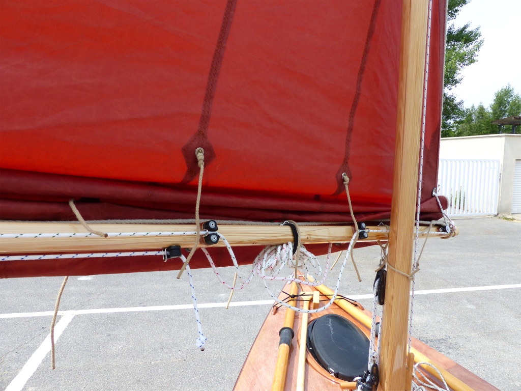 Gros plan sur la b&ocirc;me, &agrave; proximit&eacute; du m&acirc;t : de gauche &agrave; droite, on voit le coinceur de tension de bordure sous la b&ocirc;me avec le bout bleu, puis le coinceur du cot&eacute; point d'&eacute;coute de la bosse du second ris (bout vert, car coinceurs sur tribord) puis l'&eacute;lastique qui permet de ramasser le "mou" des bosses quand un ou deux ris sont pris. On a ensuite le coinceur du cot&eacute; point d'amure de la bosse du second ris, le palan d'amure et la poulie de renvoi de la bosse du second ris, et enfin le pontet de la bosse du premier ris (bout rouge). Le bout muni d'un mousqueton inox qui entoure le m&acirc;t est pris dans l'oeillet du point d'amure sur la voile et remplace l'encornat de b&ocirc;me. 