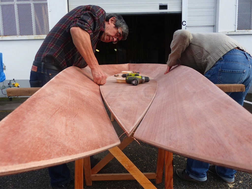 D&eacute;marrage de la couture des panneaux de coque : Pierre et Damien lient les bord&eacute;s 1 par des sutures (petits segments de fil de cuivre de 8 cm de longueur environ) de part et d'autre de la sole, le panneau de fond du Skerry. Au cours des pr&eacute;parations &agrave; cette construction, j'ai r&eacute;alis&eacute; un petit biseau &agrave; mi-&eacute;paisseur sur toutes les extr&eacute;mit&eacute;s des virures, comme on le voit ici au premier plan. La diff&eacute;rence de teinte sur la coupe est due &agrave; la couche d'&eacute;poxy que j'ai pass&eacute;e puis ponc&eacute;e sur toutes les surfaces de toutes les pi&egrave;ces. 