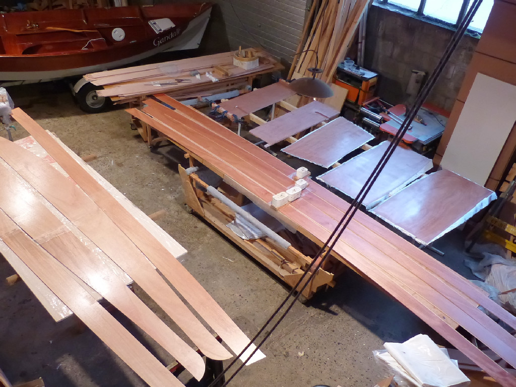 Au premier plan, ce sont des panneaux de coque de kayak Wood Duck. Sur la table centrale, on colle des demi-virure du Silmaril. Les demi-virures sont pourvues d'un marquage sur leur bord haut, à proximité du joint-puzzle : 0 boule, 1 boule, etc. Un autre marquage est présent sur le petit bord de l'extrémité arrière. Ces deux marques et le schéma fourni dans la documentation permettent d'identifier avec certitude chacune des virures. Les "boules" présentes dur le bord haut des virures seront éliminées à la ponceuse à bande ou au rabot avant de les coudre ensemble : c'est une bonne idée des marquer chaque virure dans la feuillure, car cette marque sera invisible même si on vernit tout le bateau. 