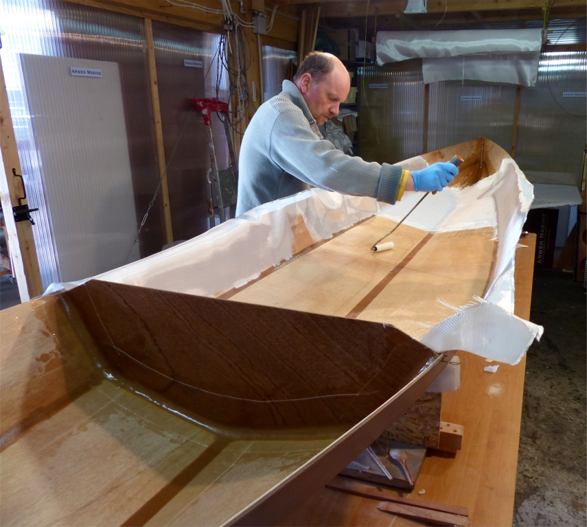 Nous arrachons les bandes Gordini et laissons un peu durcir les joints avant d'enchainer avec la stratification de la zone du cockpit. En effet, les lisières des bandes de fibre de verre que nous utilisons dans le compartiment arrière (au premier plan à gauche) et dans la pointe avant seraient gênantes dans le cockpit. Jérôme utilise un rouleau pour saturer le tissu de verre d'époxy. 