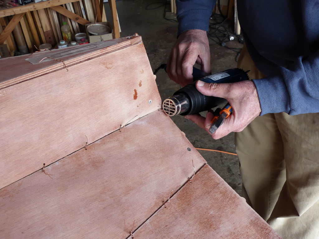 Nous commençons par extraire les sutures du pourtour du tableau, car la colle tient maintenant les extrémités des virures. Il est encore relativement facile d'extraire les sutures en les chauffant à l'aide du décapeur thermique car l'époxy est dure mais elle est encore loin de sa "maturité" maximum. La chaleur du décapeur est diffusée le long du fil de cuivre du fait de sa conductivité. Cette chaleur ramollit l'époxy qui enveloppe le fil dans le joint de colle et permet de l'extraire en tirant à la pince. Attention d'orienter le flux d'air chaud parallèlement à la surface du contreplaqué afin d'éviter de risquer de brûler la surface. 