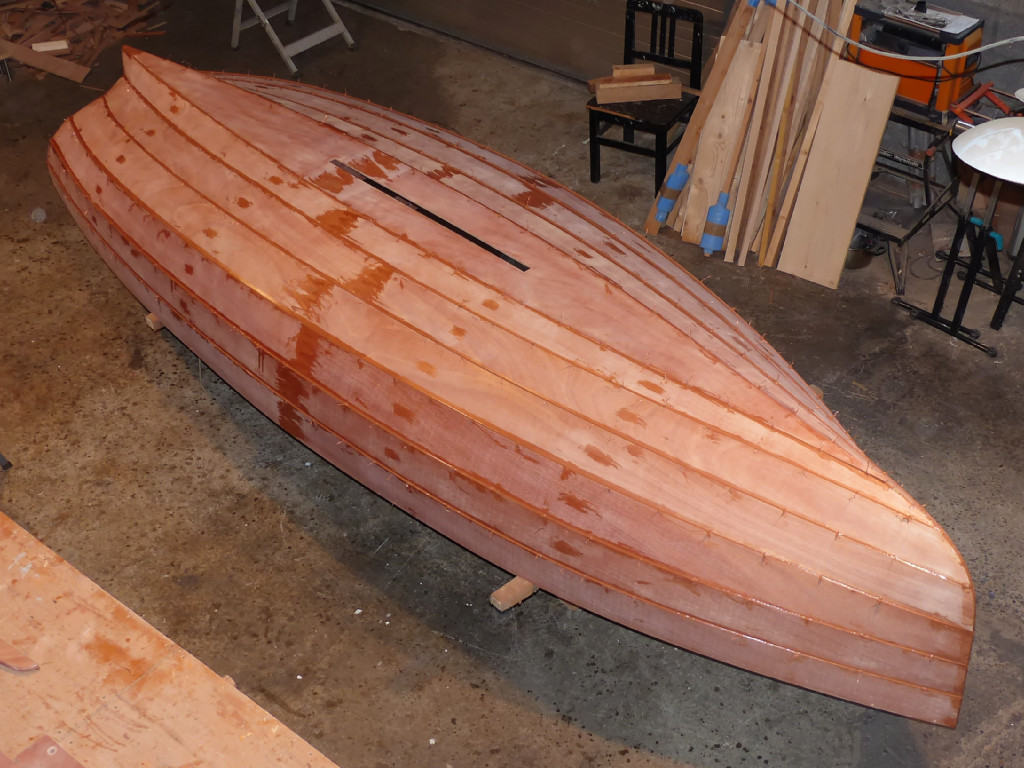 L'époxy fraîchement ajoutée apparait en "mouillé" sur la surface dépolie lors du ponçage que nous avons fait après l'imprégnation époxy générale de toutes les pièces. Nous laissons durcir ce collage pendant la nuit et le lendemain nous nous glissons sous la coque afin de couper toutes les sutures le long des joints. Nous extrayons ensuite toutes les sutures en les chauffant si besoin au décapeur thermique. Comme déjà écrit plus haut, attention d'orienter le flux d'air chaud parallèlement à la surface du contreplaqué afin d'éviter de risquer de brûler la surface. 