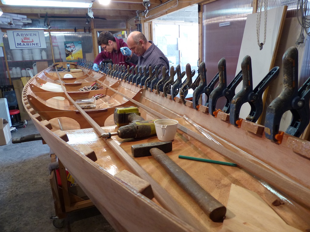 Le vendredi 14 avril, Olivier et Hugo (en rouge bordeaux) sont revenus comme pr&eacute;vu, de m&ecirc;me que Pierre, (devant Hugo) qui doit commencer la construction accompagn&eacute;e de son Skerry le lendemain. Notez que les Pinces sont maintenant &agrave; c&ocirc;t&eacute; des cales, coll&eacute;es de la veille, car nous collons le liston en premier. On voit que Hugo est en train d'encoller chacune des cales &agrave; l'&eacute;poxy charg&eacute;e, en vue du collage de la serre, en seconde phase. 