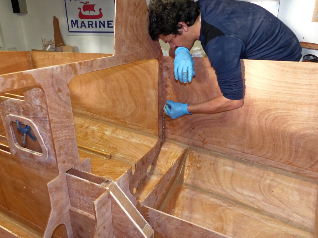 Martin utilise une seringue pour déposer un cordon d'époxy chargée autour de la cloison qui termine le petit rouf. Ce cordon sera ensuite formé en arc de cercle dans l'angle, puis couvert d'un ruban de fibre de verre de 100 mm de large qui le renforcera considérablement en l'ancrant plus largement à la surface du contreplaqué.