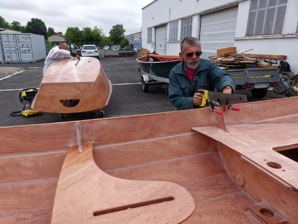 Jean-Louis atttaque son bateau à la scie égoïne. Les presses de part est d'autre servent à éviter que les listons se décollents de part et d'autre de la coupe. Une épaisseur de carton ondulé entre les deux cloisons avant nous donne un espace de 5 mm pour sécuriser la coupe. Notez que Jacques est arrivé entretemps pour terminer la construction de son GIS, on le voit occupé à poncer à l'arrière-plan.