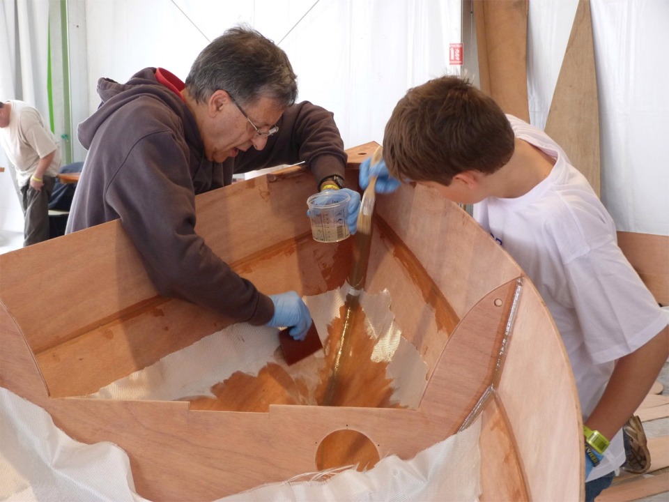 Michel et François font de même : ils versent la résine époxy sur le tissu de verre et l'étalent et la font pénêtrer à la raclette. François utilise un petit pinceau pour saturer le tissu dans les endroits peu accessibles à la raclette. 