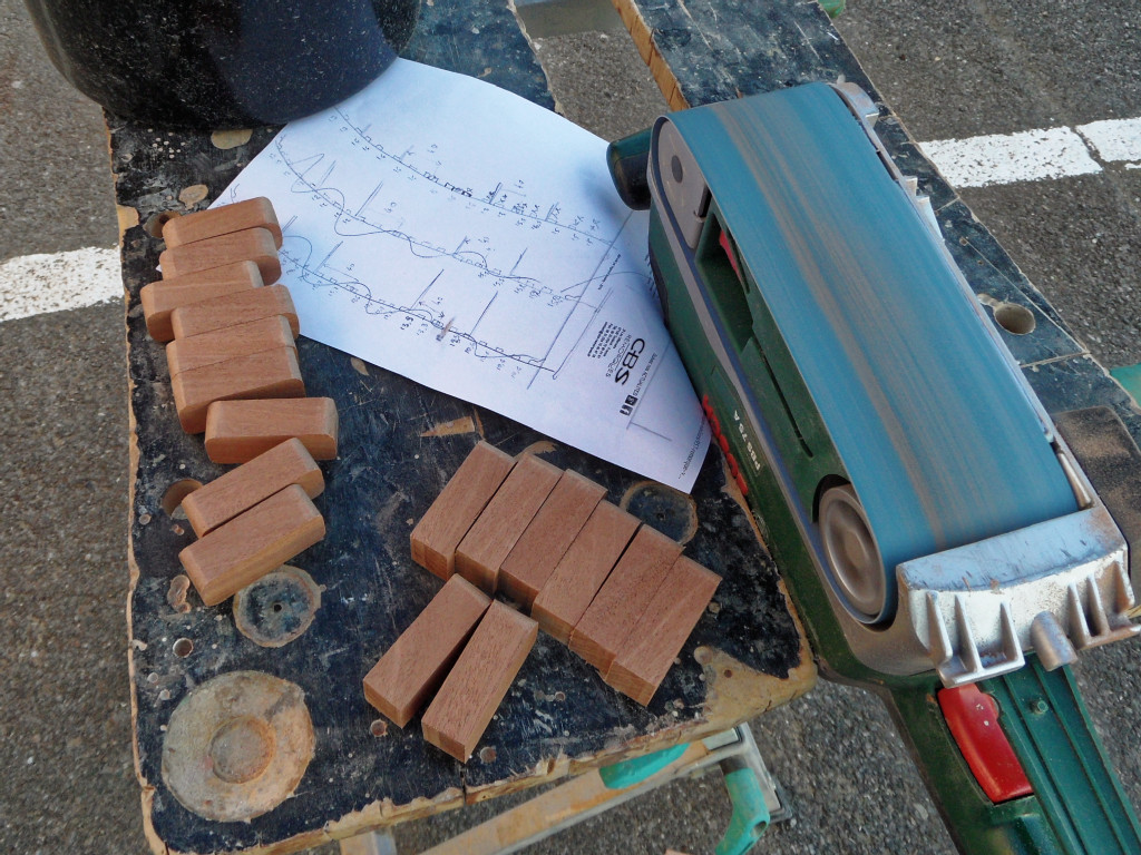 Préparation des cales de la serre ajourée : l'une des 5 lattes de sapelli est découpée en segments de 50 mm de longueur, et les quatres petites arêtes d'une grande partie de ces cales est arrondie sur la ponceuse à bande. Sur une autre partie des cales, seules les deux petites arêtes d'une des deux extrémités sont arrondies, car ces cales seront adossées à la tête de l'une des quatre cloisons (il faut donc 16 de ces cales). 